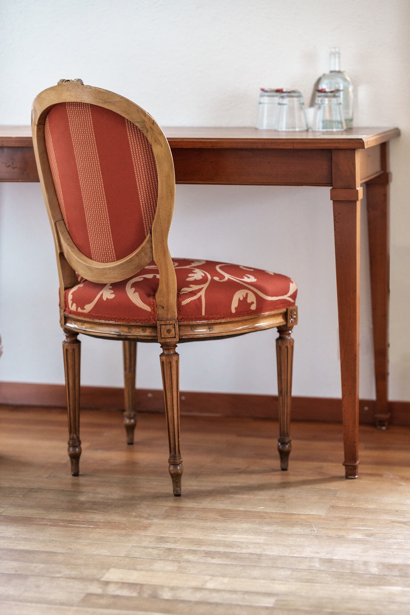 Wooden chair with red patterned upholstery stands in front of a wooden table on wood flooring.